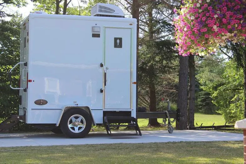 Restroom Trailer sitting on a paved area surrounded by trees and colorful flowers, featuring a door with a male symbol and an air conditioning unit on top.