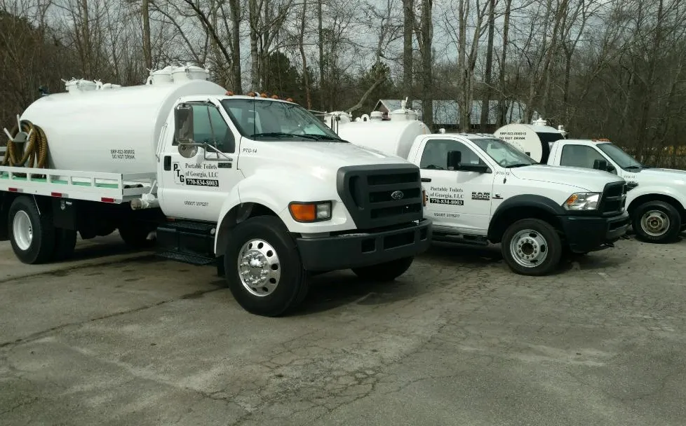 A line of three service vehicles parked on a gravel surface. The foremost vehicle is a large white truck with a tank mounted on the back, labeled for Portable Toilets. The second vehicle is a smaller white truck, while the third one is a pickup truck, both also displaying Portable Toilets alongside a phone number. In the background, bare trees are visible, typical of a late winter or early spring setting.
