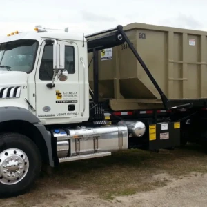 Roll Off Dumpster on a truck, showcasing a large, beige dumpster attached to the back, with the truck's silver cab and shiny chrome accents partially visible. The background is an outdoor area with grass and dirt.